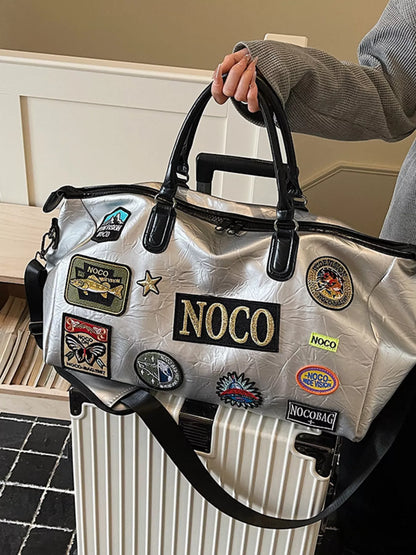 Silver bag with NOCO patches held by a person next to a suitcase.