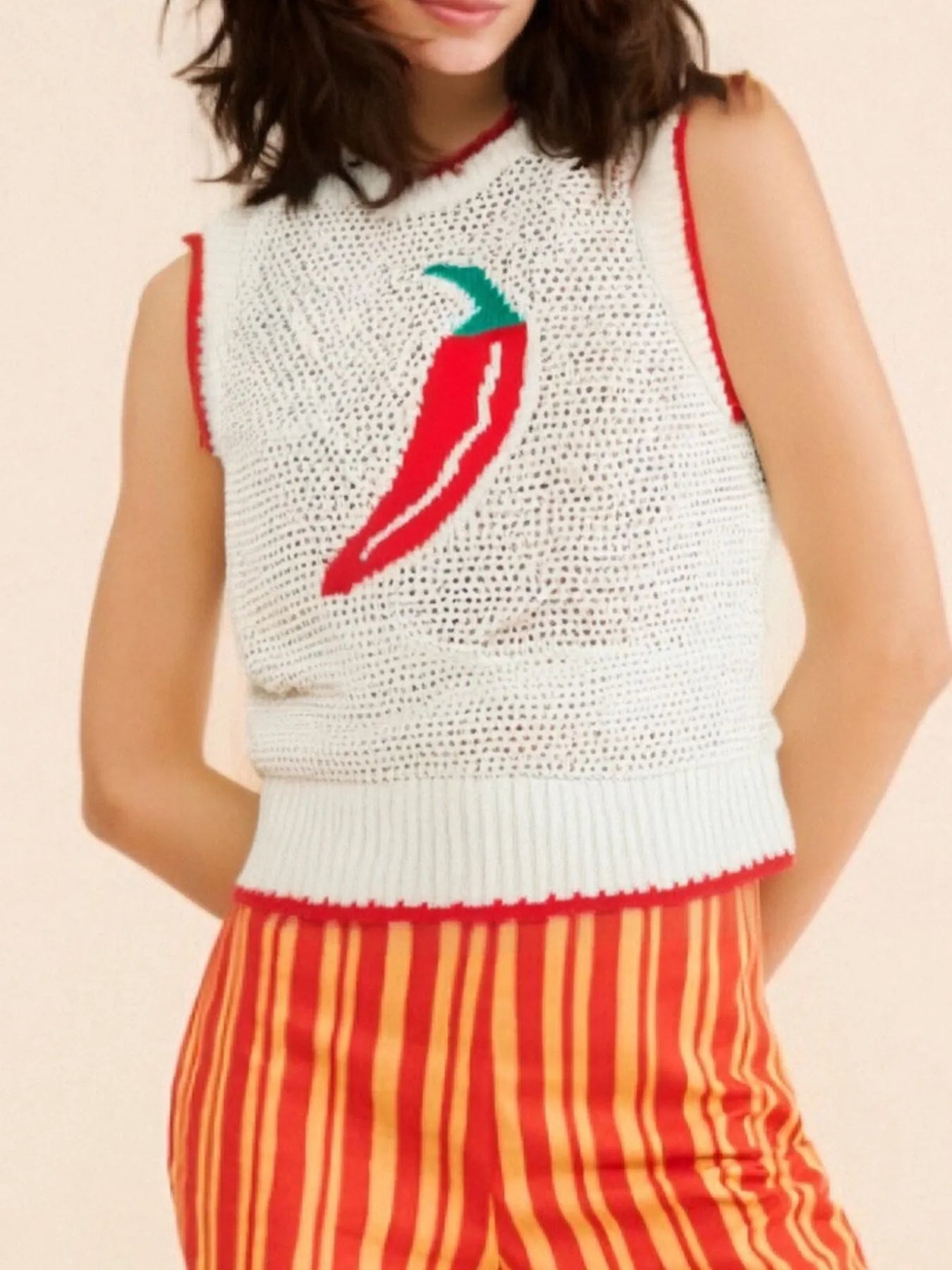 Person wearing a white sleeveless crochet top with a red chili pepper design and orange striped pants on a beige background.