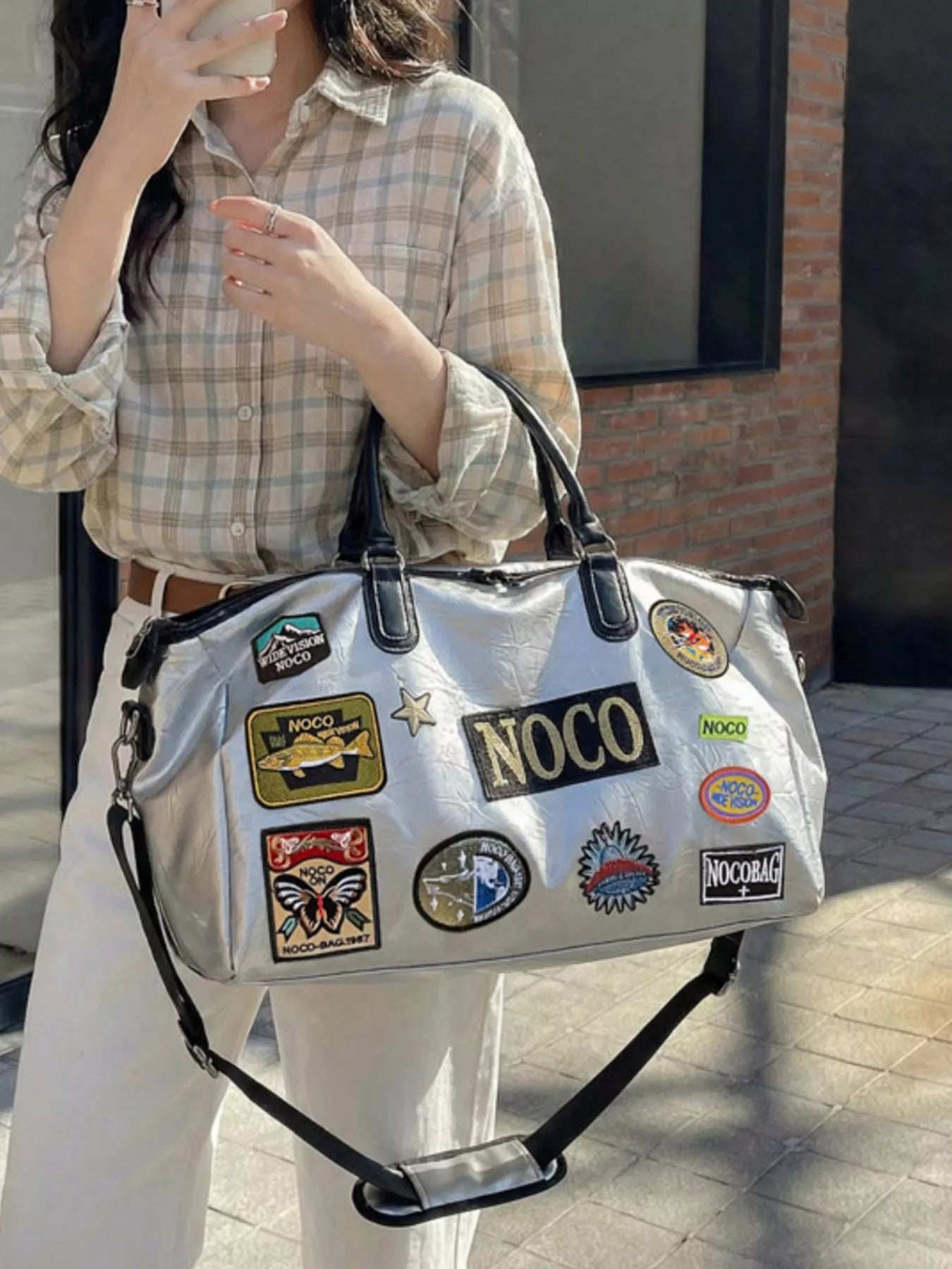 Person holding a silver bag with multiple patches, including 'NOCO', against a neutral background.