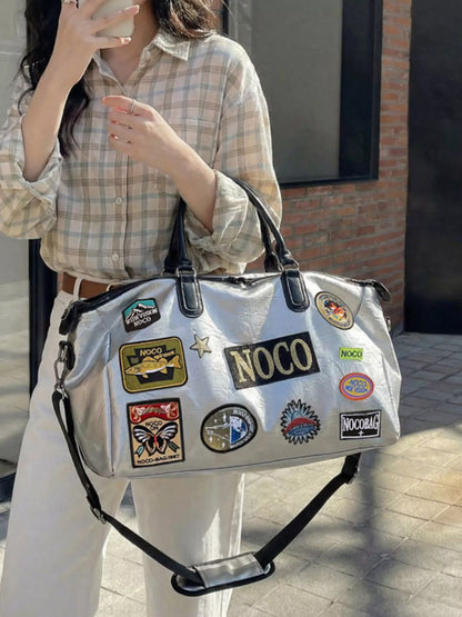 Person holding a silver bag with multiple patches, including 'NOCO', against a neutral background.