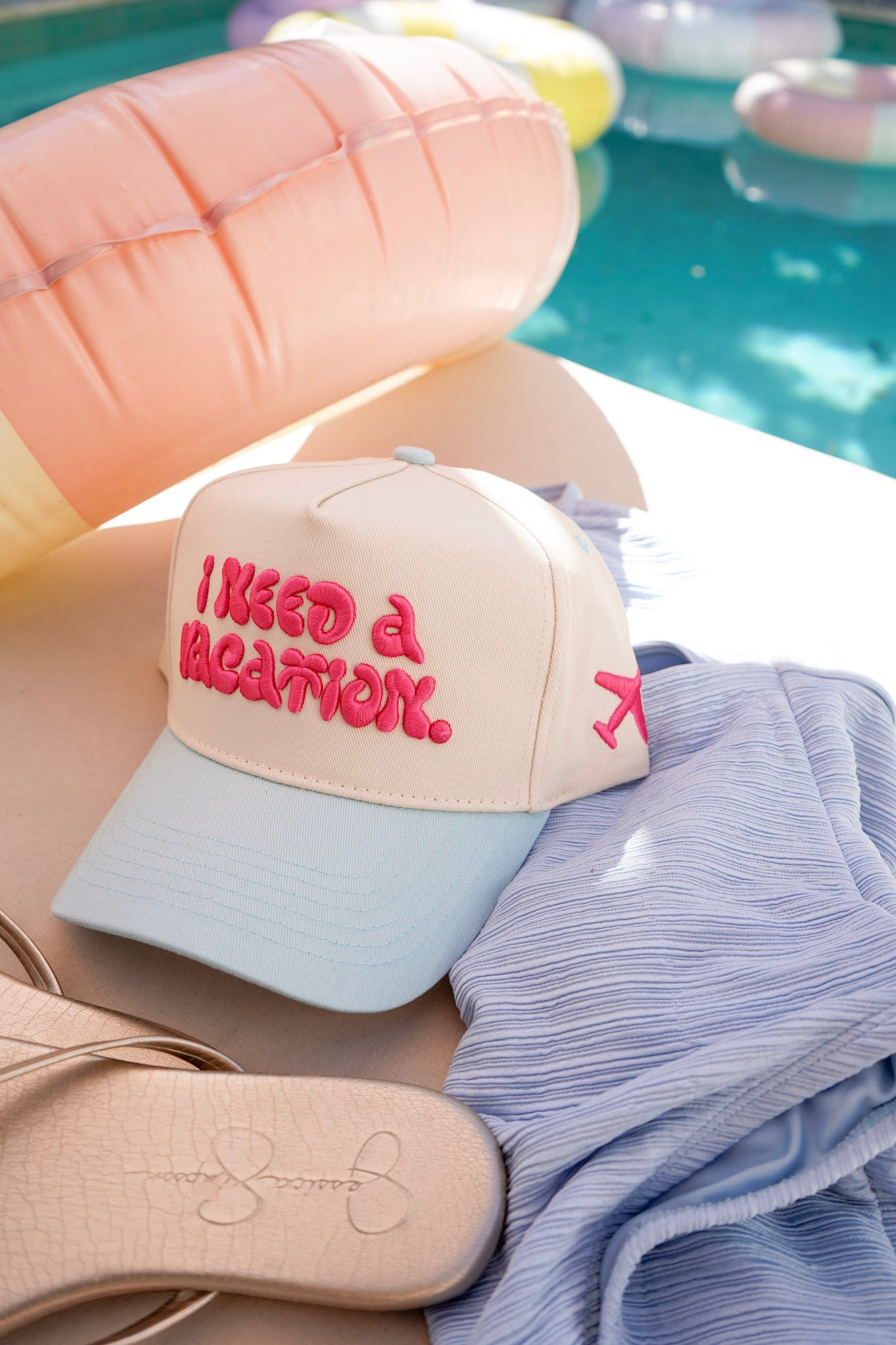 Beige cap with 'I need a vacation' text, sunglasses, and towel by a pool.
