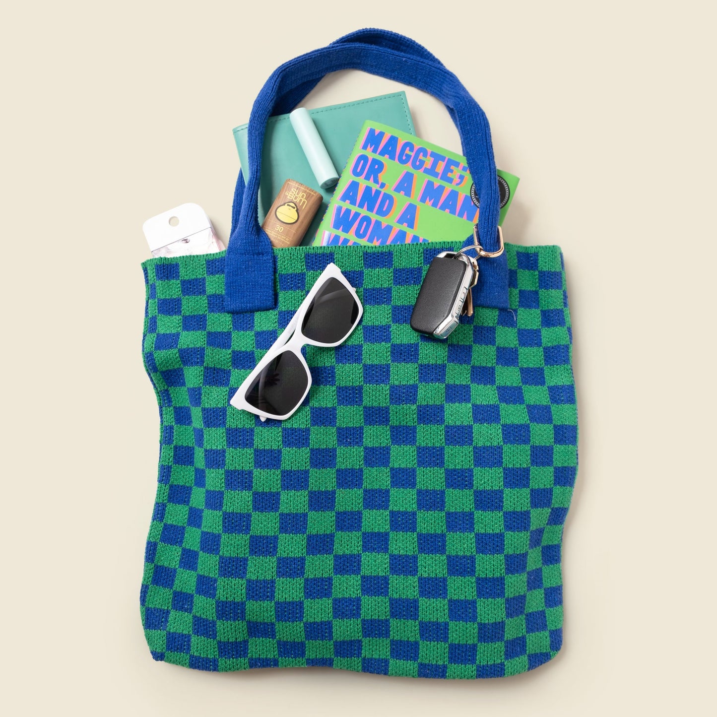 Green and blue checkered tote bag with sunglasses and books on a beige background