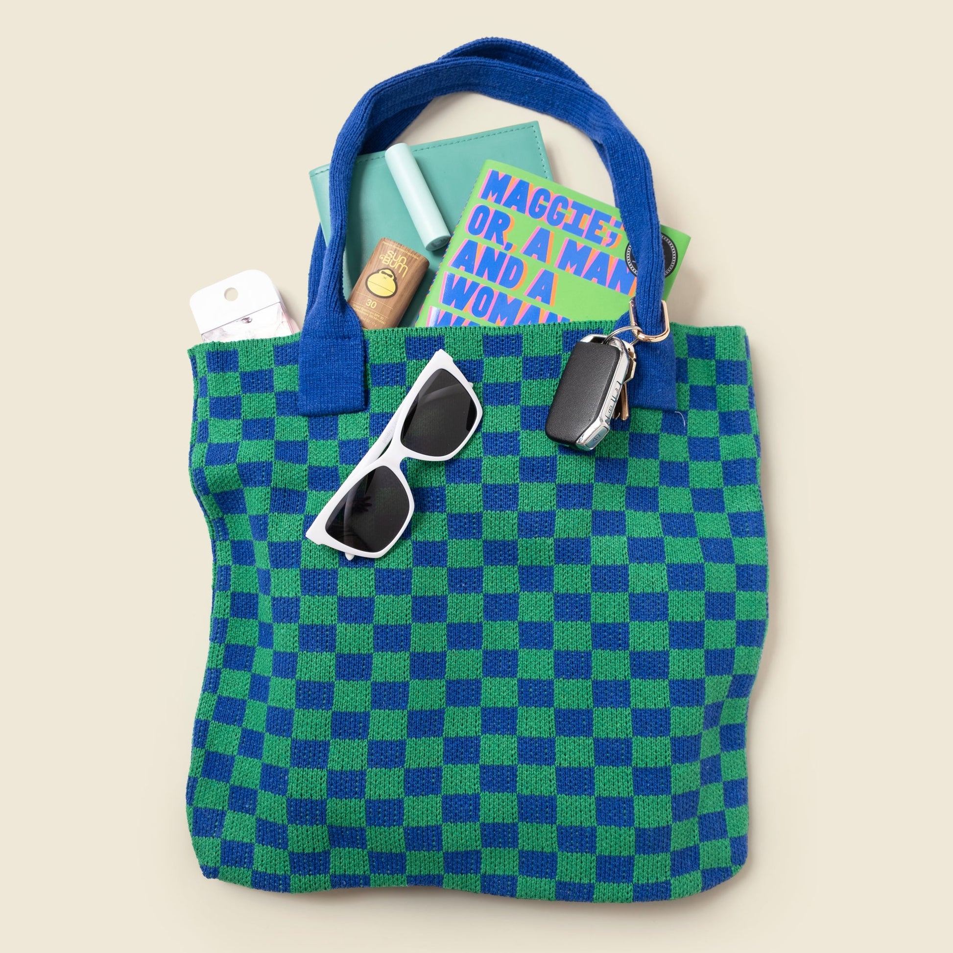 Green and blue checkered tote bag with sunglasses and books on a beige background