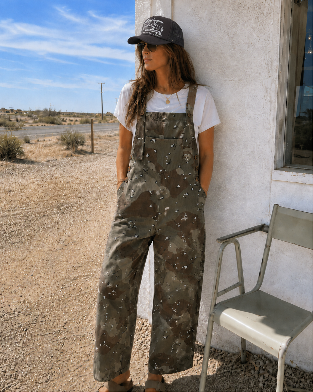 Person wearing camouflage overalls and a cap leaning against a building with a desert landscape in the background.