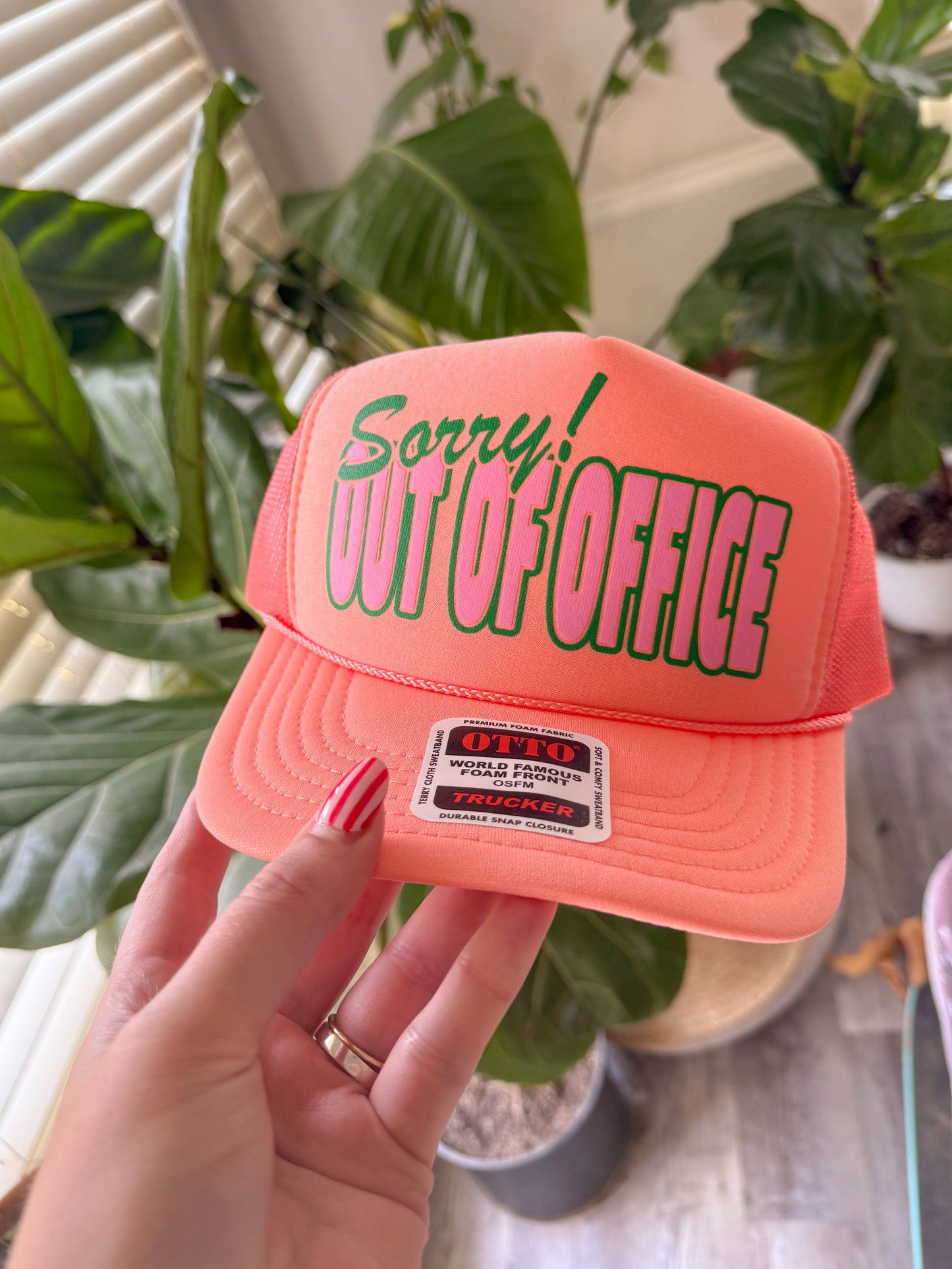 Pink cap with 'Sorry! Out of Office' text held by a hand in front of green plants.