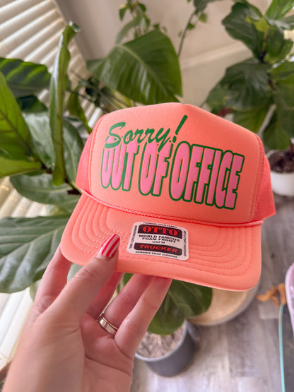 Pink cap with 'Sorry! Out of Office' text held by a hand in front of green plants.