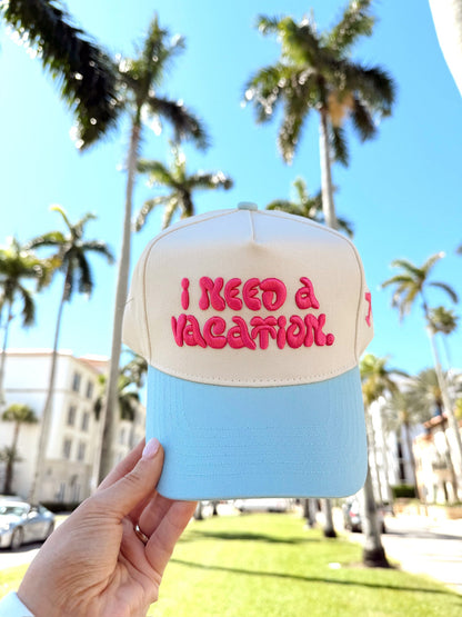 Cap with 'I Need a Vacation' text held in front of palm trees and clear blue sky