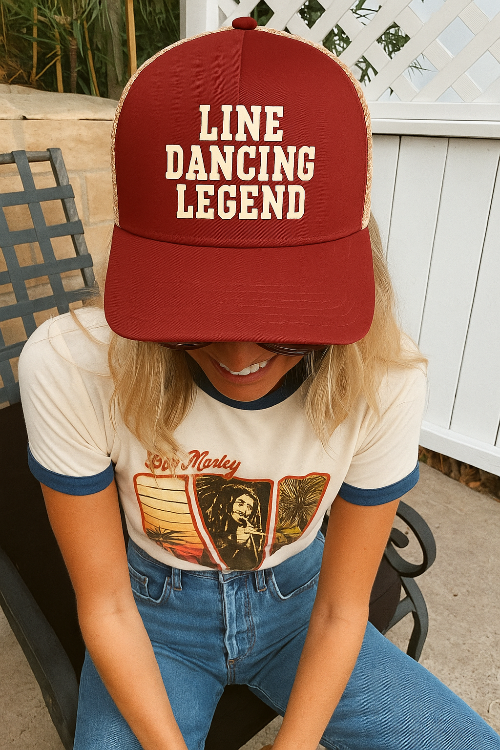 A young woman wearing the red Line Dancing Legend hat with a retro top and jeans, sitting on a patio
