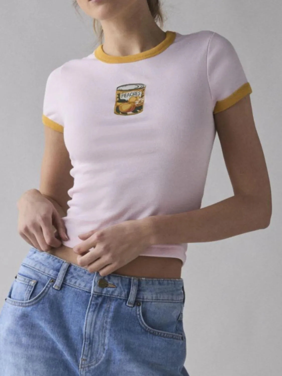 Light pink baby tee with yellow trim and retro-style canned peach graphic on the chest.