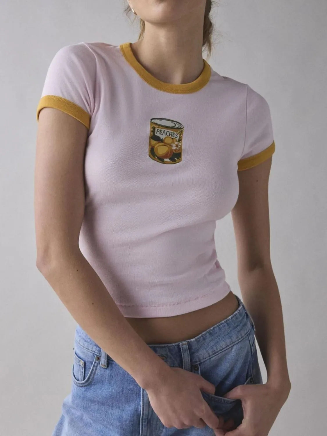 Light pink baby tee with yellow trim and retro-style canned peach graphic on the chest.