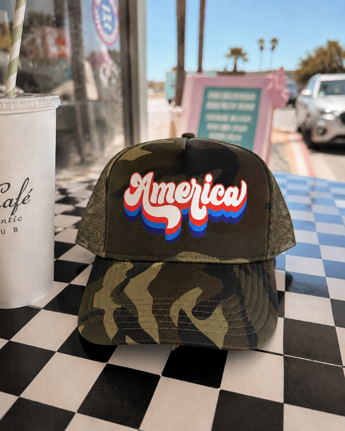 Camouflage cap with 'America' text on a checkered table, with a drink and blurred background.