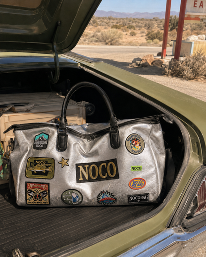 Silver duffel bag with NOCO patches in a car trunk with desert landscape in the background