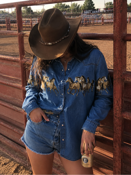 Person wearing a denim shirt with horse prints and a cowboy hat, standing in front of a metal fence.