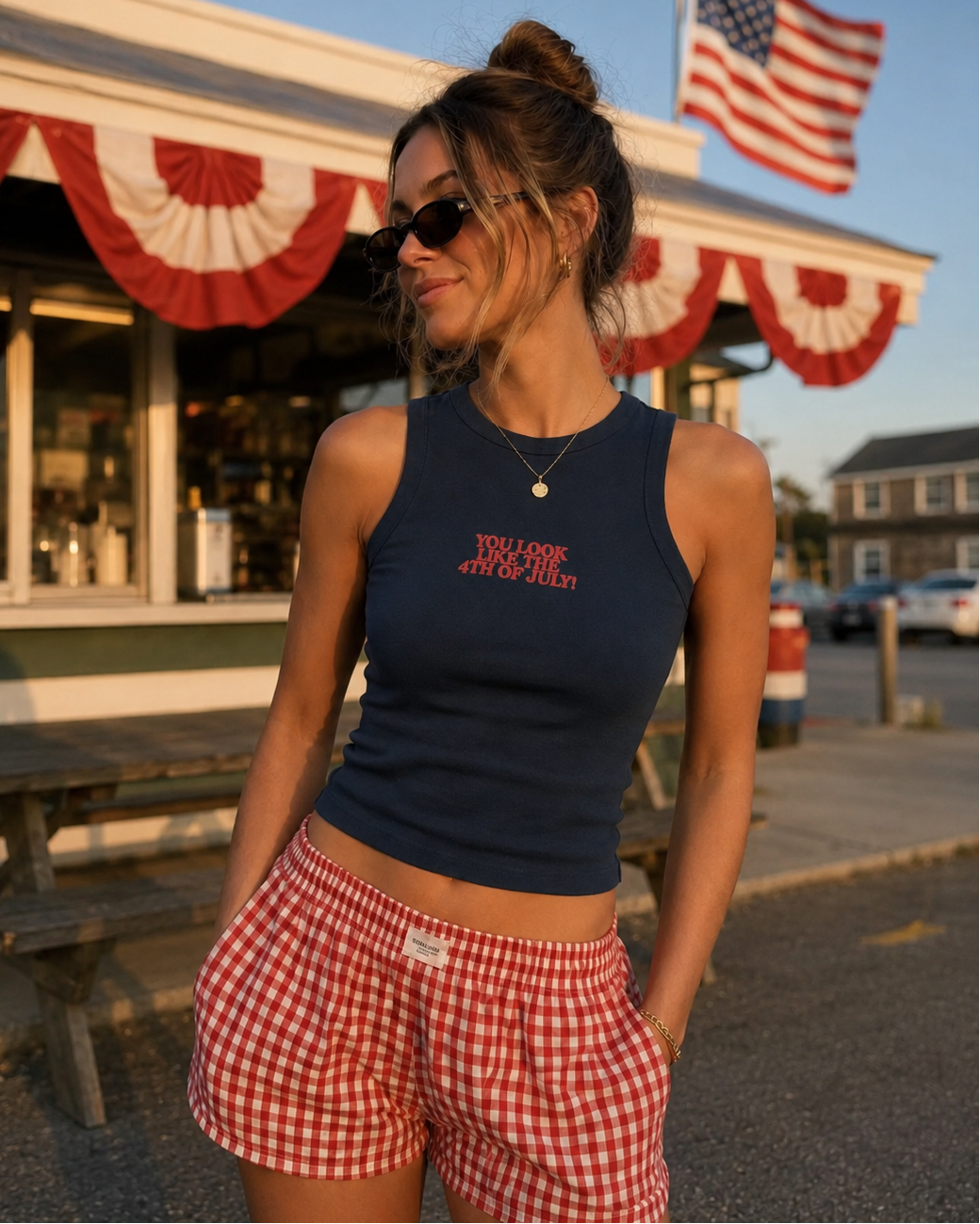 Woman wearing a navy tank top with text and red checkered shorts standing in front of a building with an American flag.