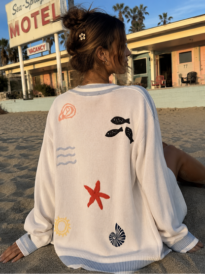 Person wearing a white sweater with beach-themed designs sitting on a sandy beach in front of a motel.