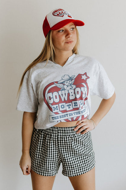 Person wearing a white t-shirt with 'Cowboy Motel' design, black and white checkered shorts, and red and white cap.