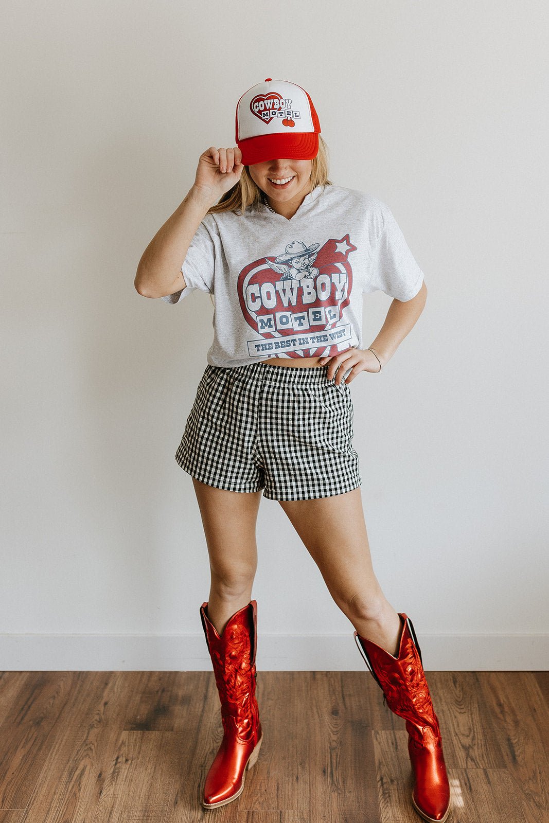 Person wearing a white t-shirt with a graphic design, plaid shorts, and red cowboy boots, standing against a plain background.