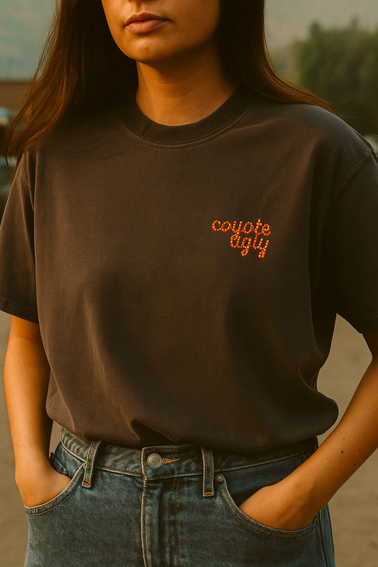 Woman wearing a charcoal graphic tee that says “coyote ugly” in marquee-style letters, styled with high-rise jeans for a vintage western look.