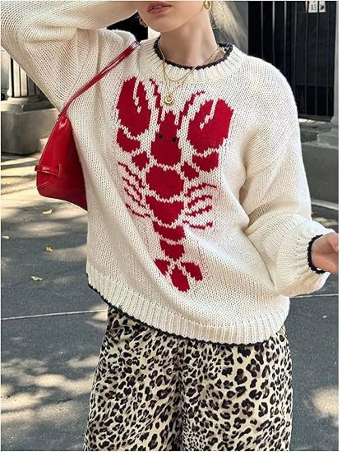 Person wearing a white sweater with a red abstract design, holding a red bag on a street.