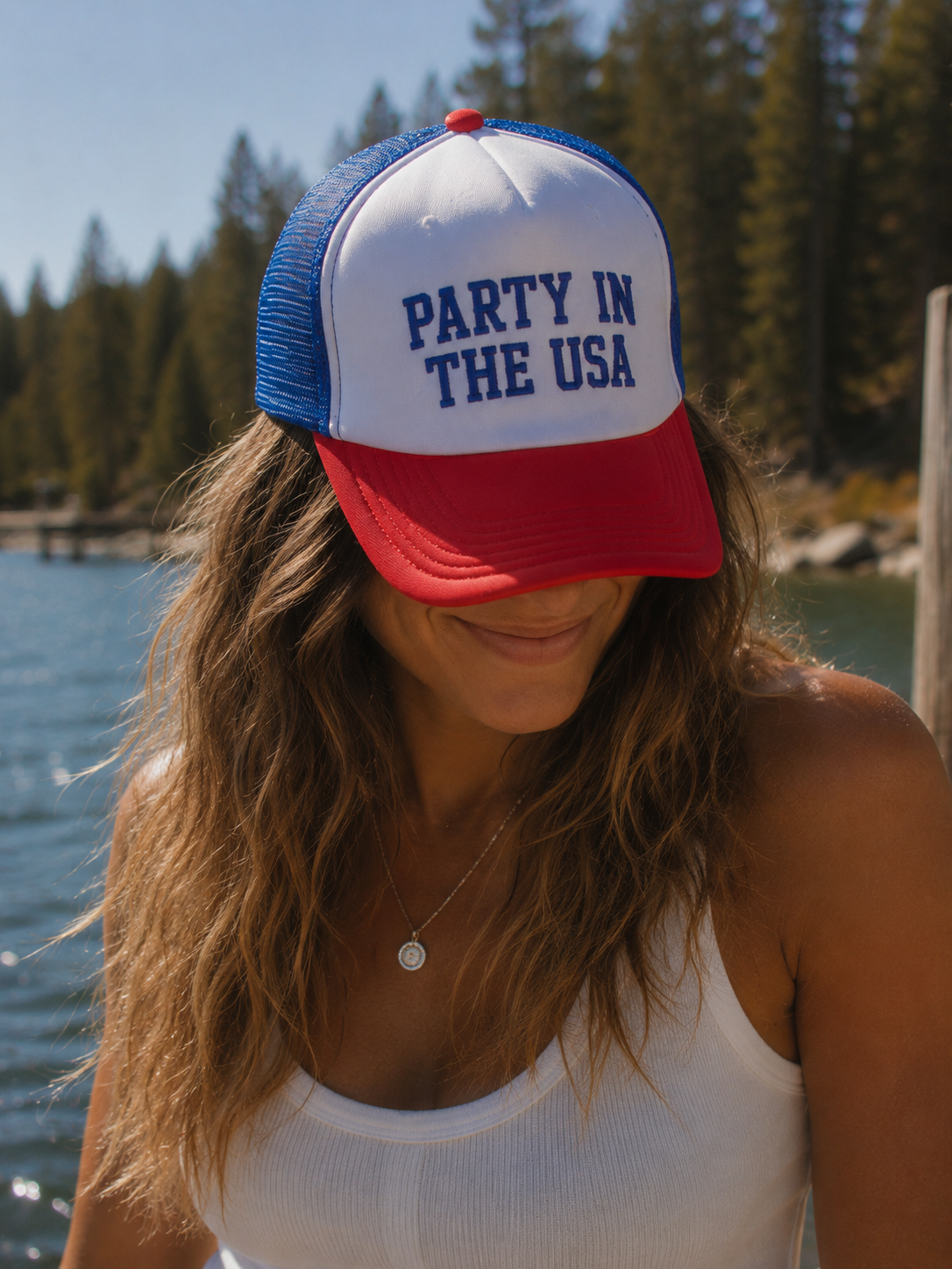Person wearing a 'Party in the USA' cap by a lake with trees in the background