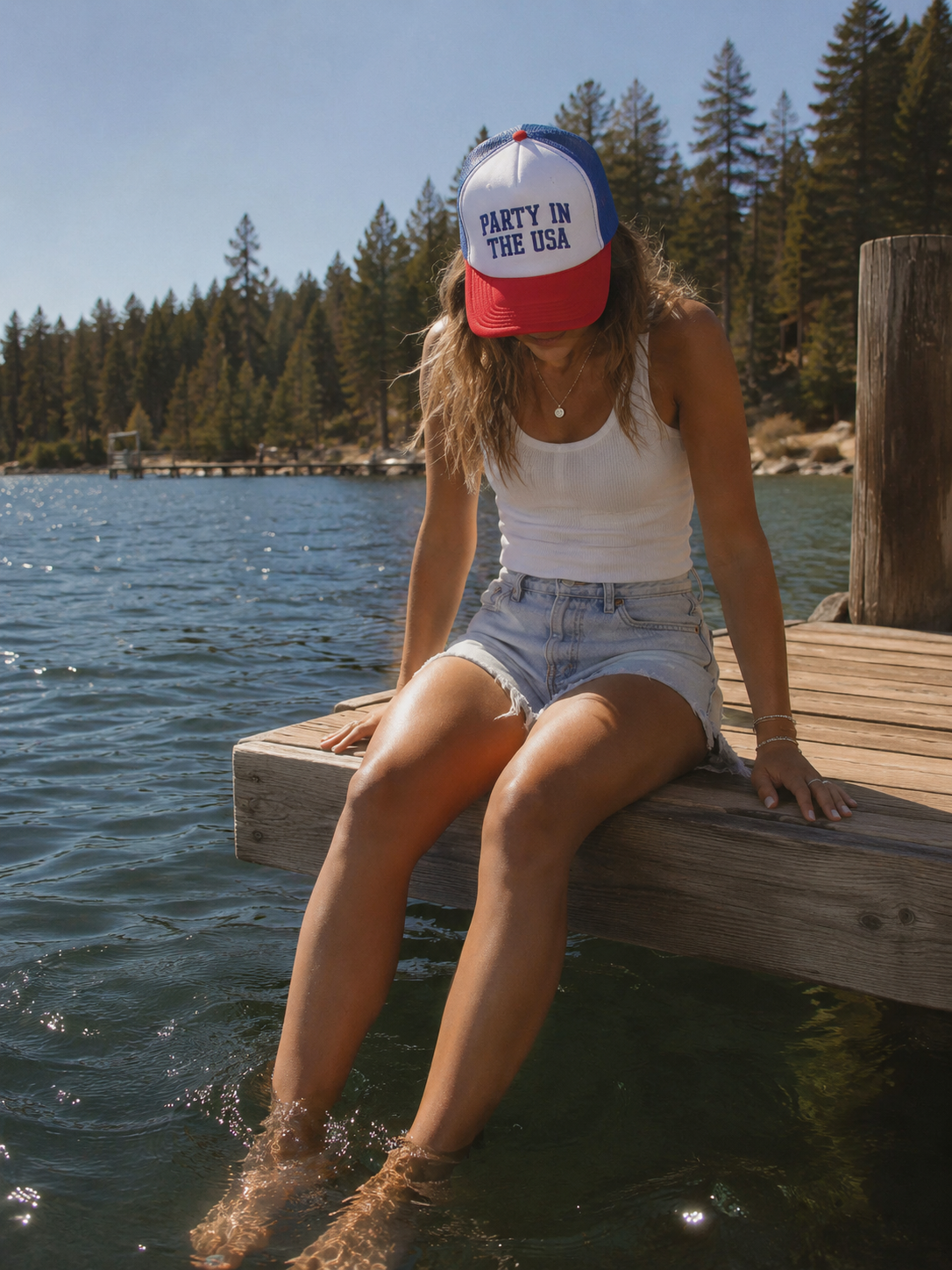 Person sitting on a dock by a lake wearing a 'Party in the USA' cap.
