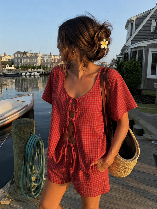 Woman in a red checkered outfit standing on a dock by a waterfront with houses and boats in the background.