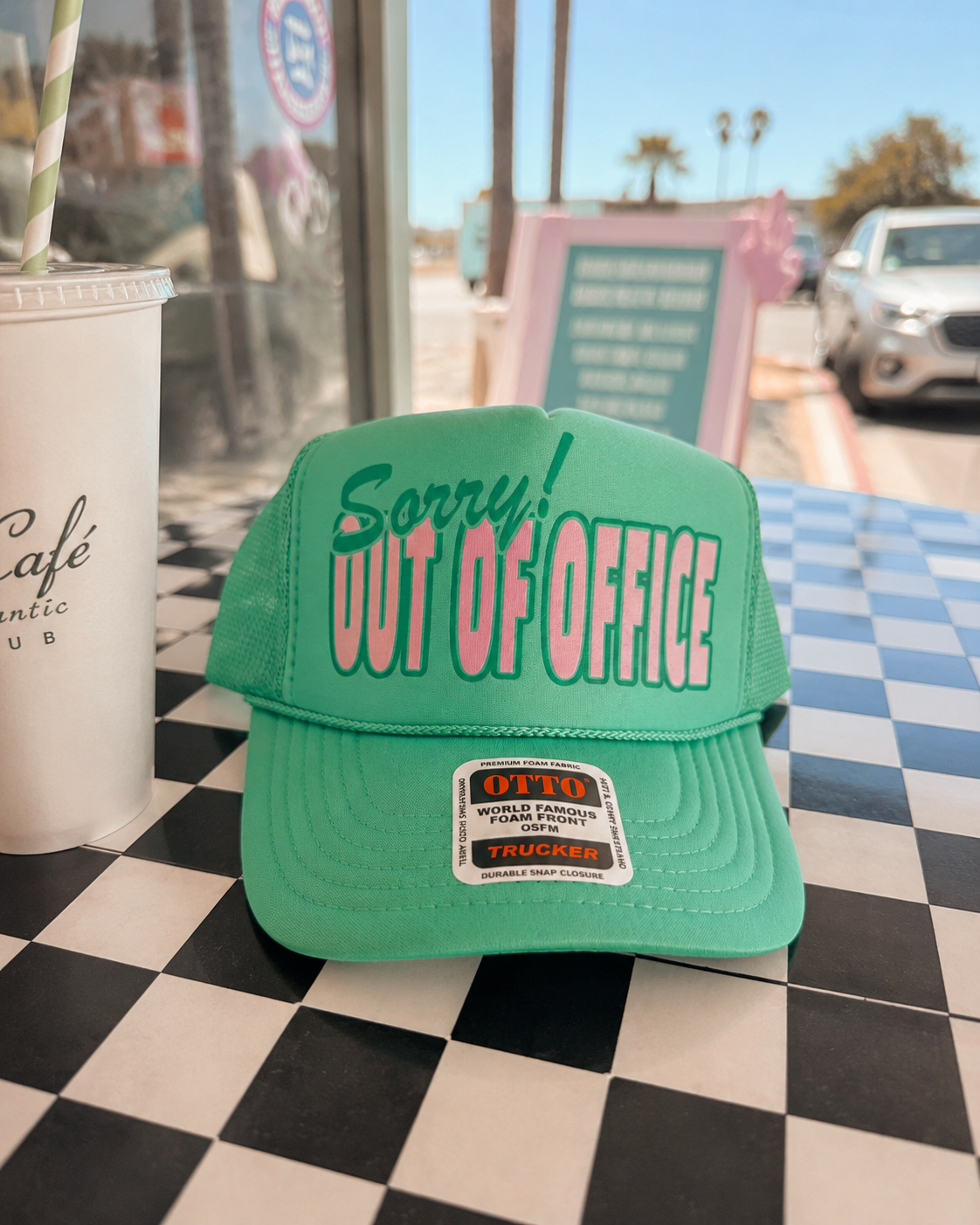 Green cap with 'Sorry! Out of Office' text on a checkered table with a cup and straw.