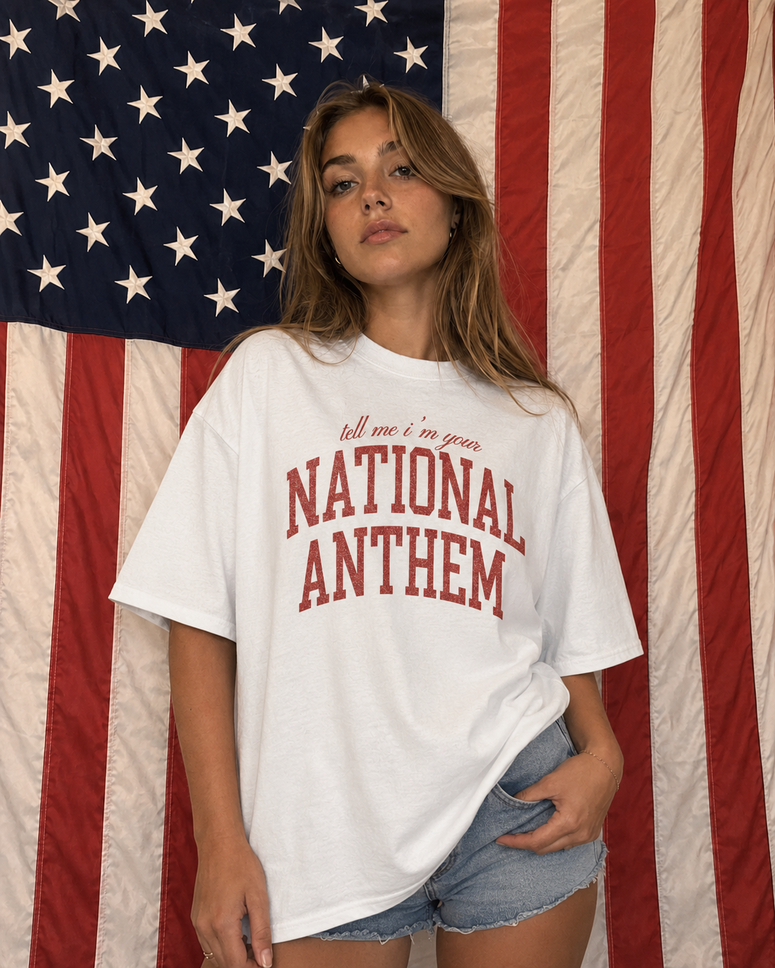 Person wearing a white t-shirt with 'tell me i'm your NATIONAL ANTHEM' text in front of an American flag.